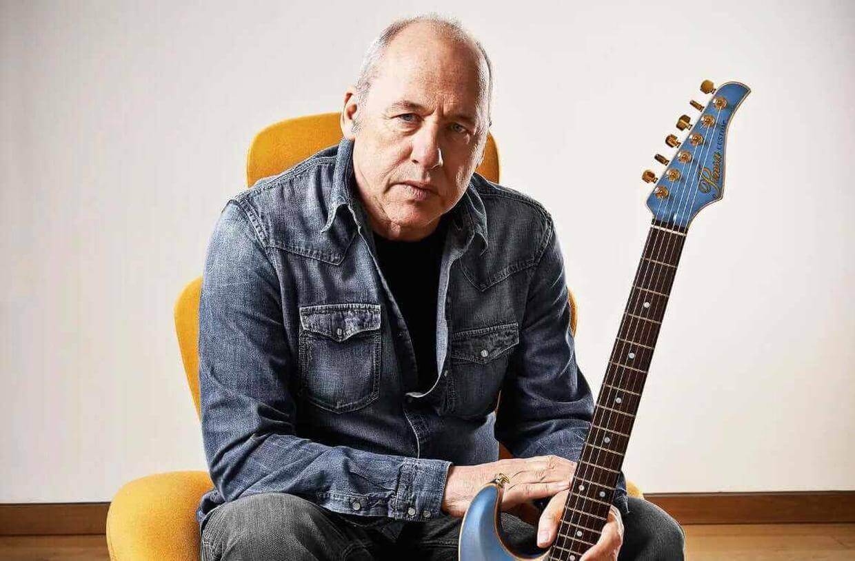 A musician holding a guitar, illustrating how to play guitar like Mark Knopfler, featuring his signature style.