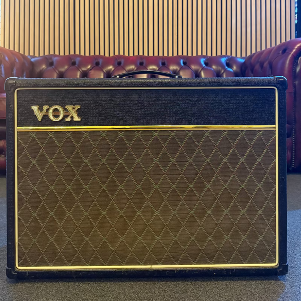 A Vox guitar amplifier with a black front, gold trim, and brown diamond-patterned grille cloth, positioned in front of a red leather tufted sofa and a slatted wood wall.