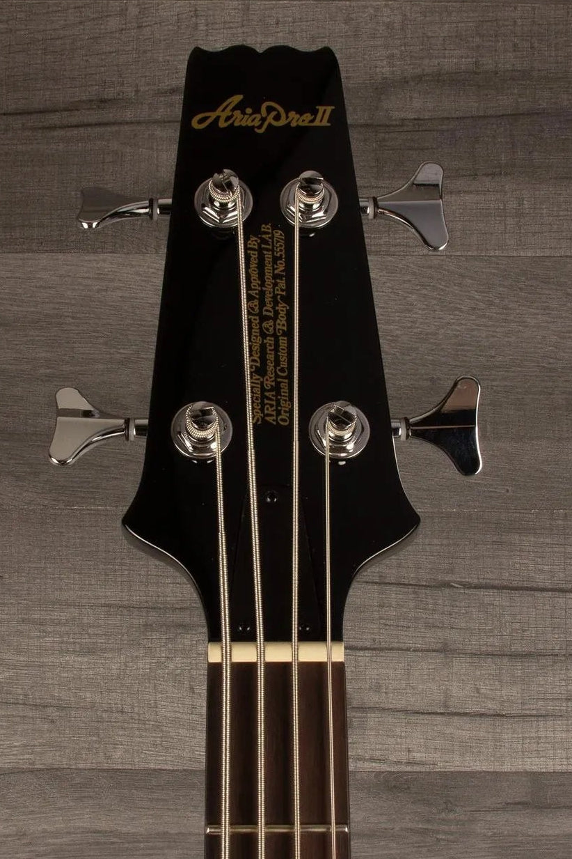 Close-up of a black Fender bass guitar headstock with four tuning pegs and gold lettering, set against a gray wood plank background.