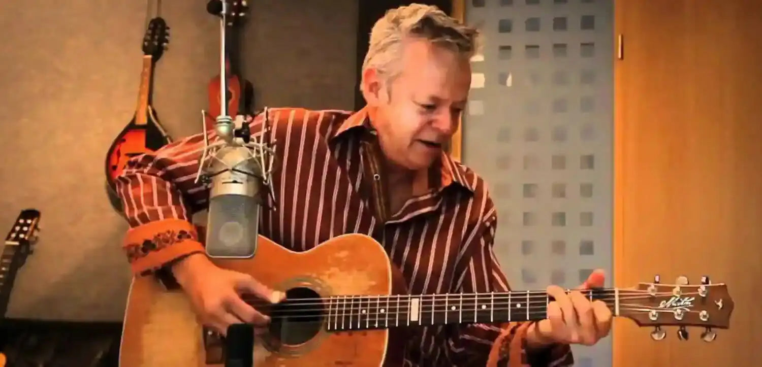Man playing an acoustic guitar in a recording studio with microphones and other instruments.